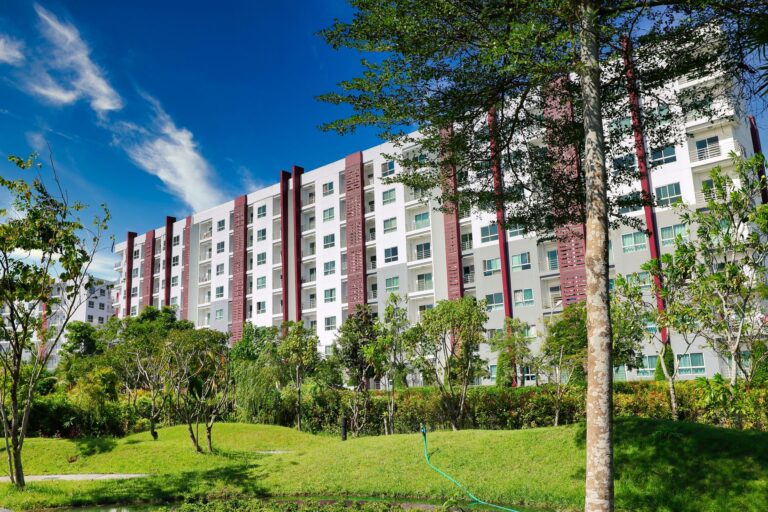 A mid-rise apartment building with a shared lawn, hedges and young trees in front